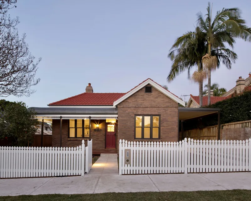 A brick house with a red tiled roof and a white picket fence at dusk